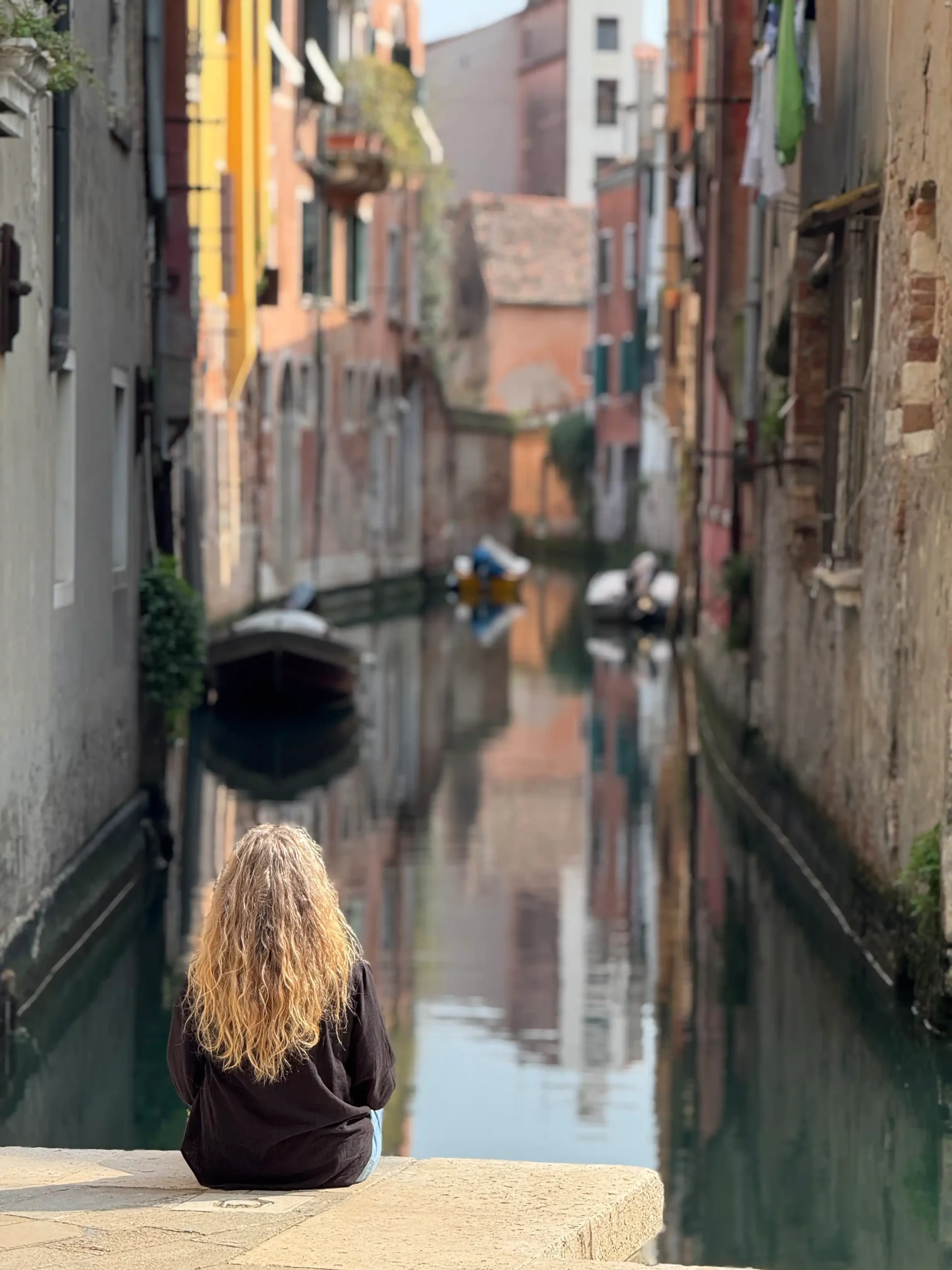 Moteris sėdi ant tilto turėklų ir žvelgia į siaurą Venecijos kanalą su valtimis