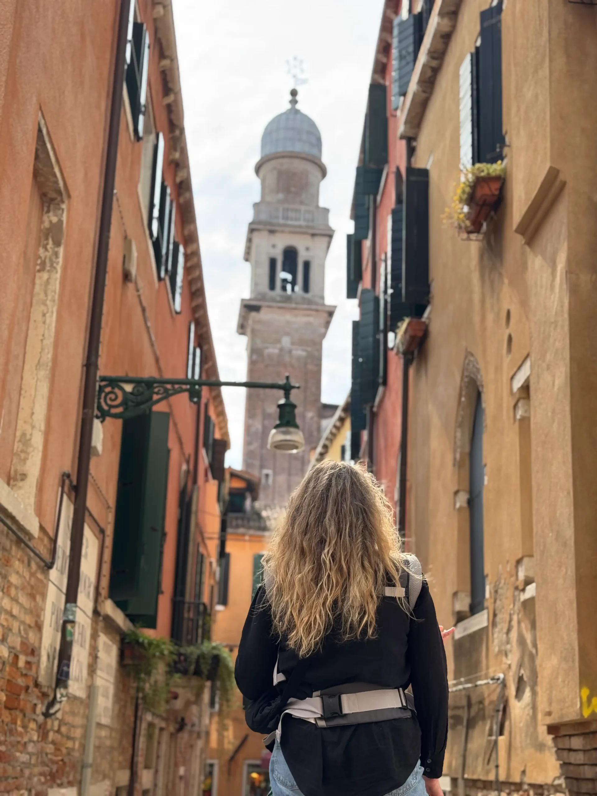 Moteris eina siaura Venecijos gatve, fone matomas bažnyčios kupolinis bokštas