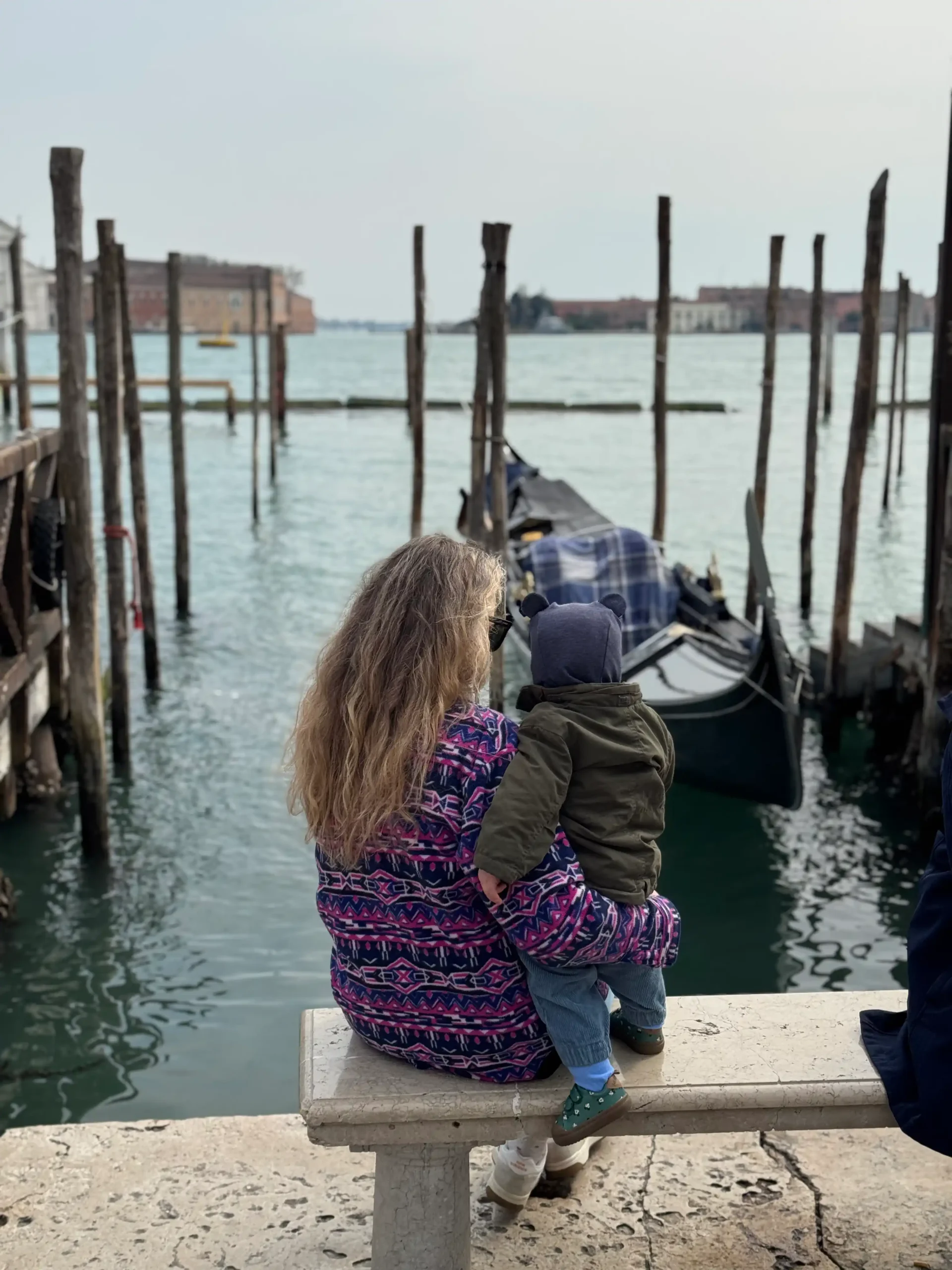Mama su mažu vaiku sėdi ant suoliuko ir žiūri į gondolas Venecijos lagūnoje