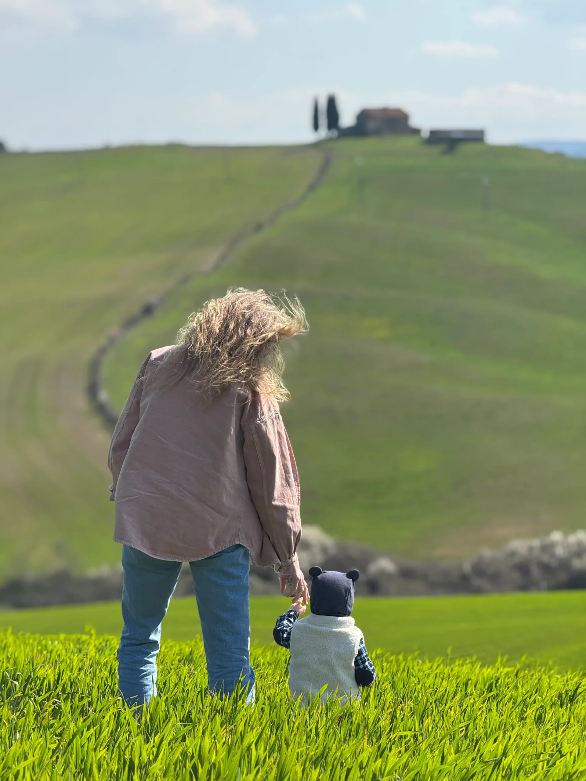Toskana su kūdikiu: 4 naktys Chianti regione (patirtis 2026) Val d'Orcia su vaiku — mama ir kūdikis žaliuose pavasario laukuose, fone Cappella Vitaleta