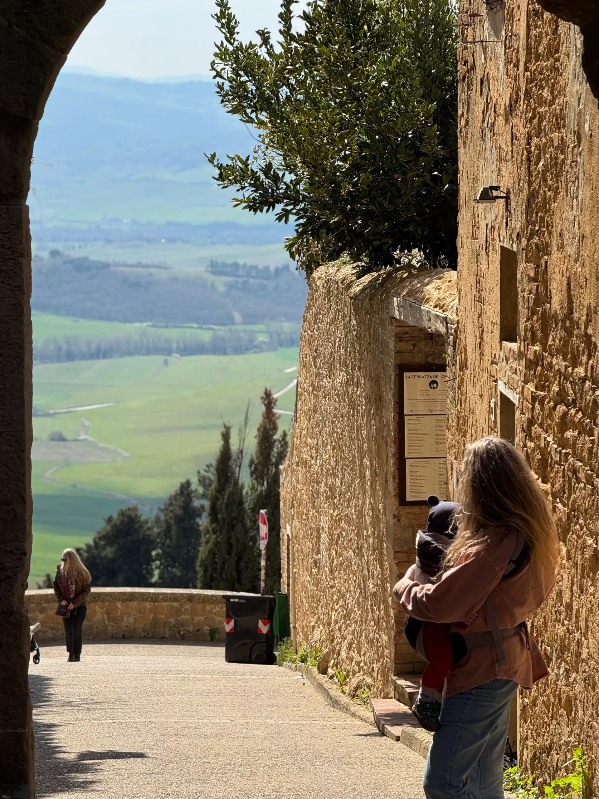 Toskana su kūdikiu: 4 naktys Chianti regione (patirtis 2026) Pienza — mama su kūdikiu ant rankų miestelio vartų arkoje, fone atsiveria Val d'Orcia slėnio panorama