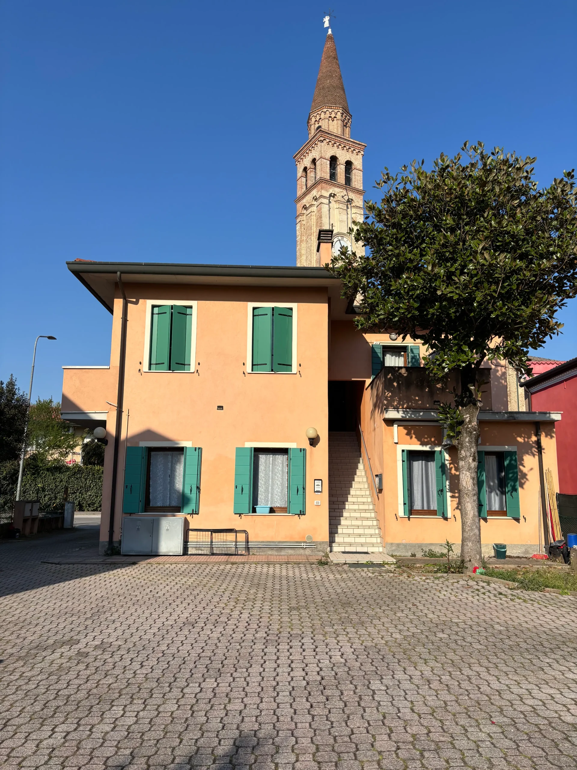 Itališkas kaimas su gelsvų sienų namu žaliomis langinėmis ir bažnyčios bokštu mėlyno dangaus fone