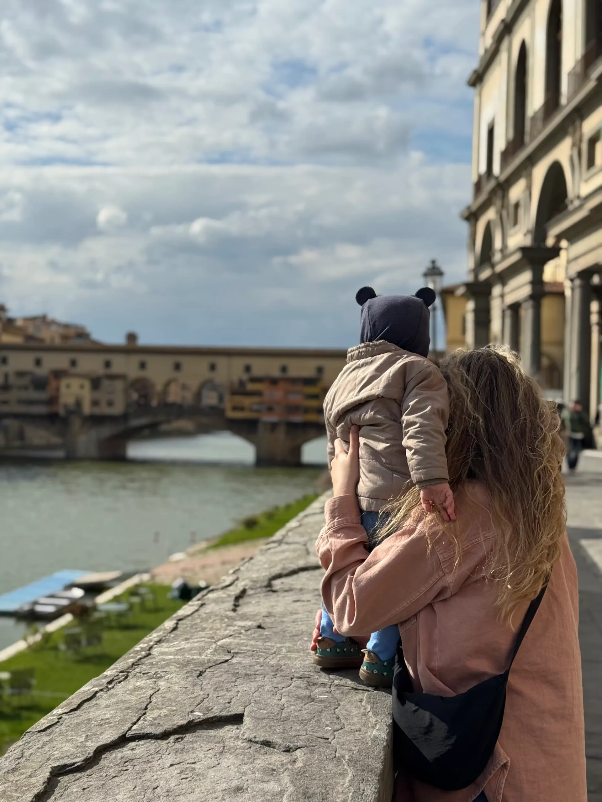 Toskana su kūdikiu: 4 naktys Chianti regione (patirtis 2026) Mama su mažu vaiku ant rankų žiūri į Ponte Vecchio tiltą per Arno upę Florencijoje