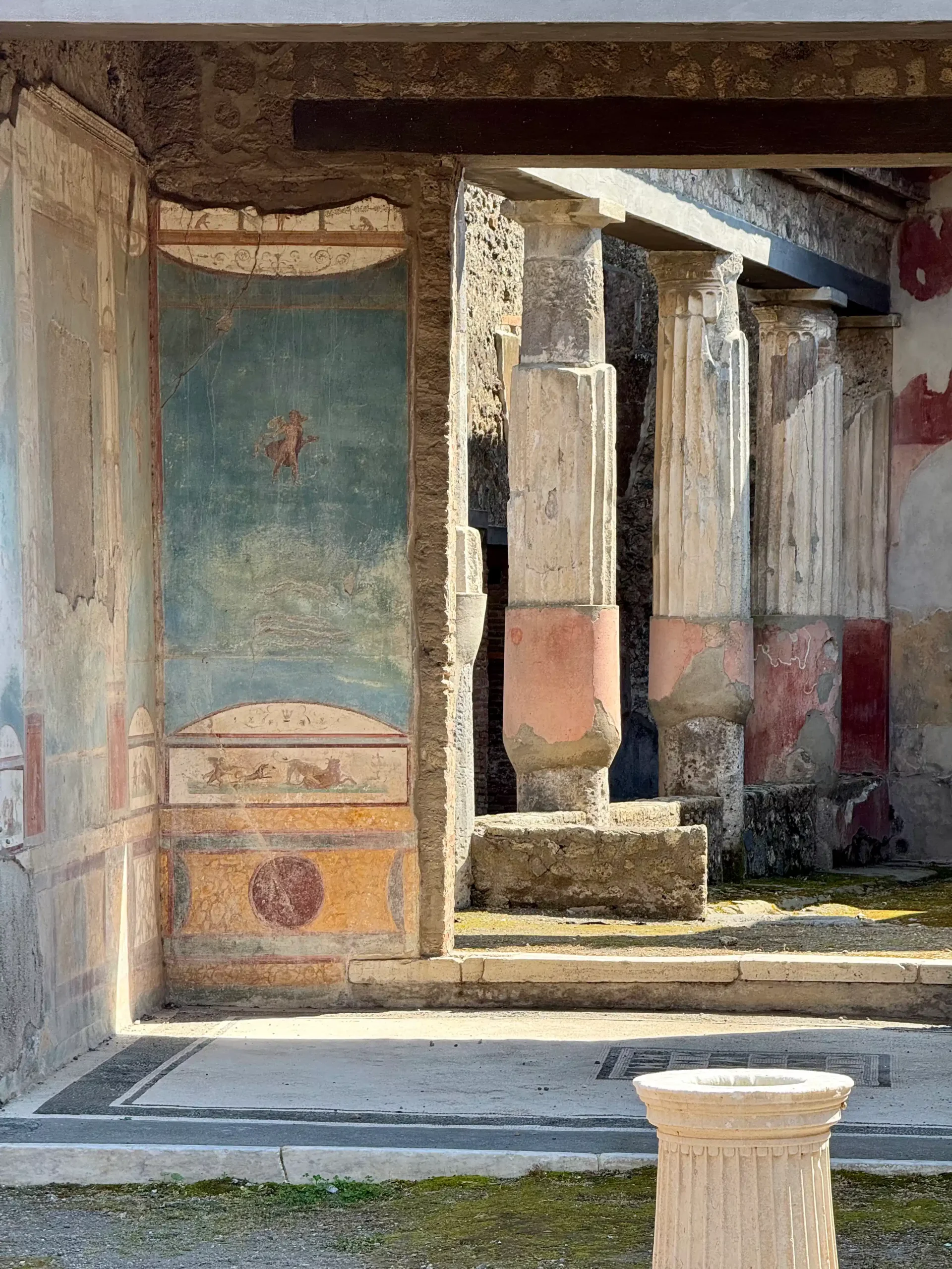Gerai išlikusios romėniškos freskos Pompėjos name: mėlono fono mitologinė scena, kolonu portikinis koridorius ir marmurinis fontano postamentas