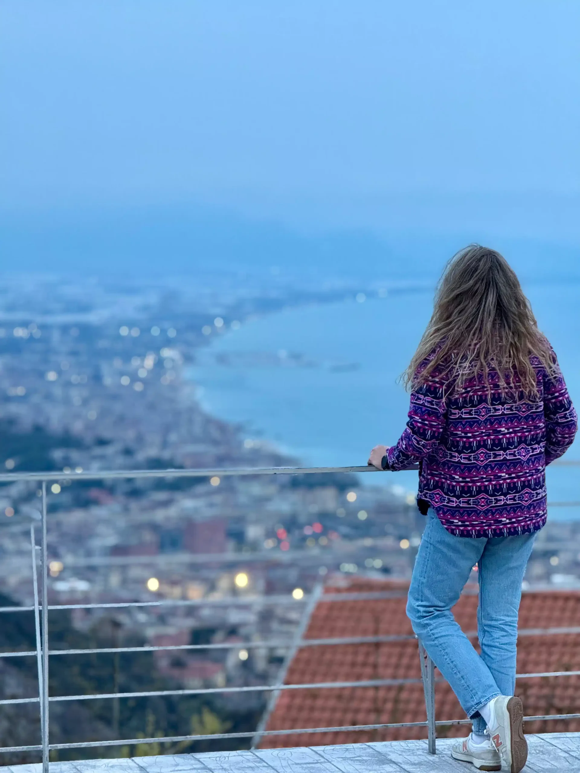 Moteris ryškiu megztuku stovi ant aukštos terasos ir žvelgia į pakrantės miestą bei įlanką sutemų mėlynoje