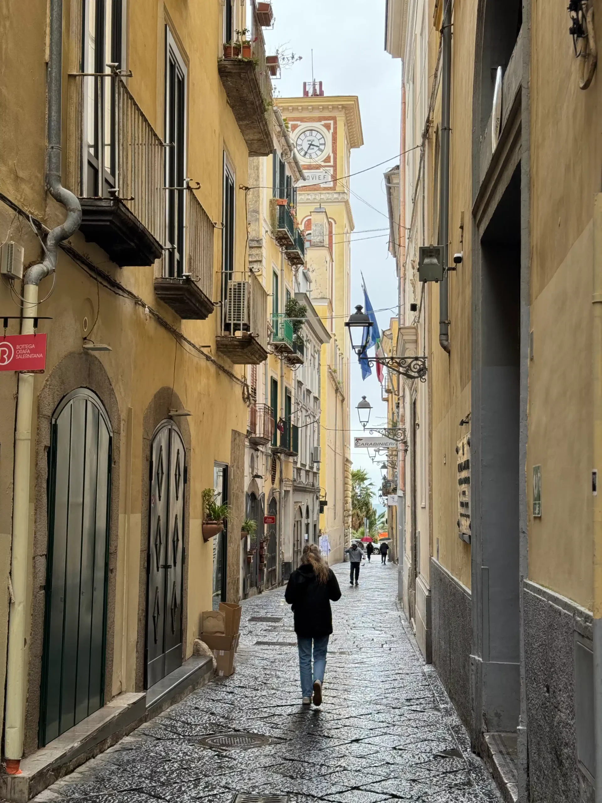 Moteris eina siaura lietingu grįstiniu gatve italų senamiesty; tolumoje matomas laikrodžio bokštas ir Italijos bei ES vėliavos