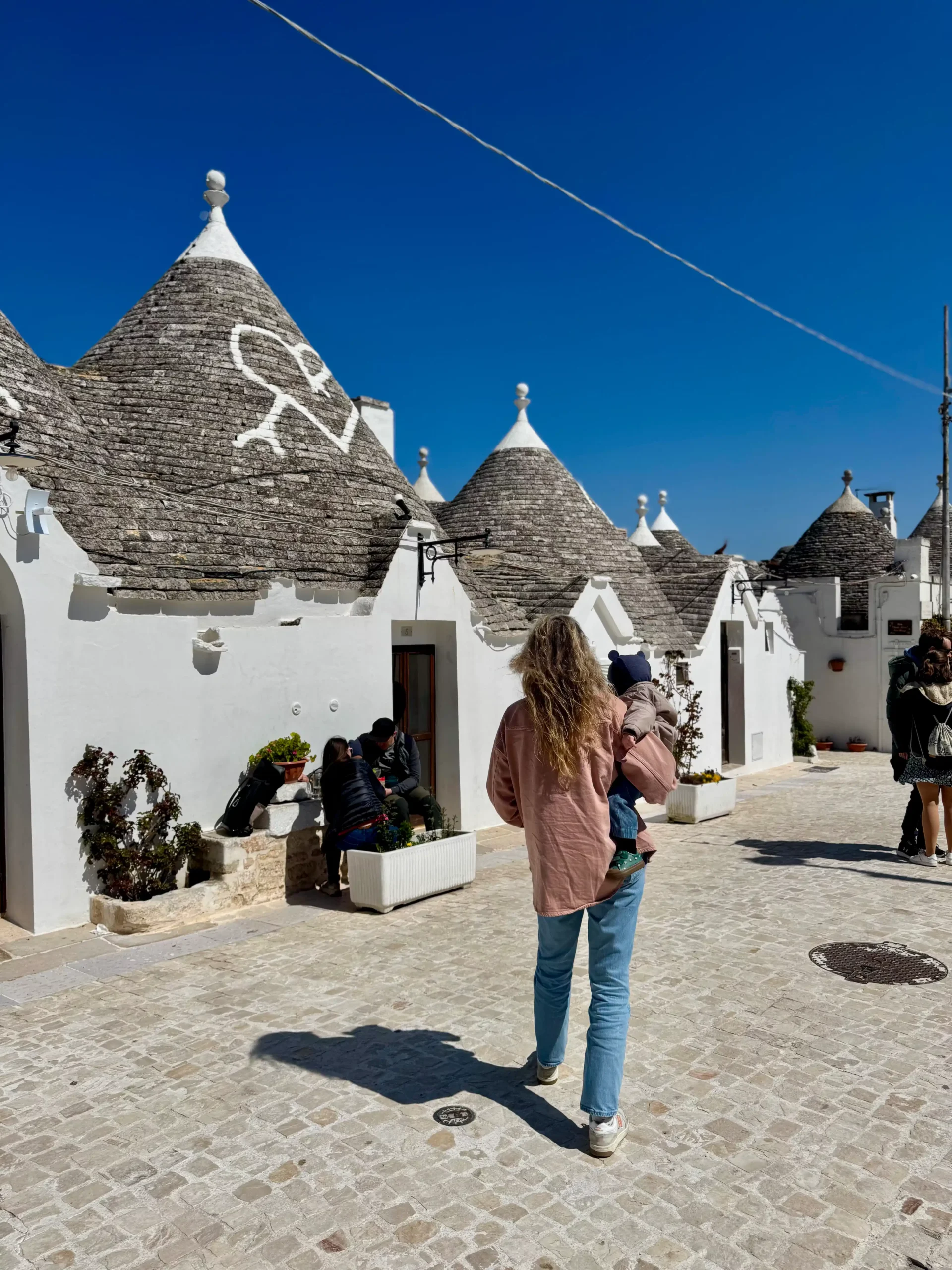 Moteris su kūdikiu ant rankų eina akmenimis grįstu kiemu tarp baltų trulli namelių su kūginiais akmeniniais stogais Alberobello mieste, Italija