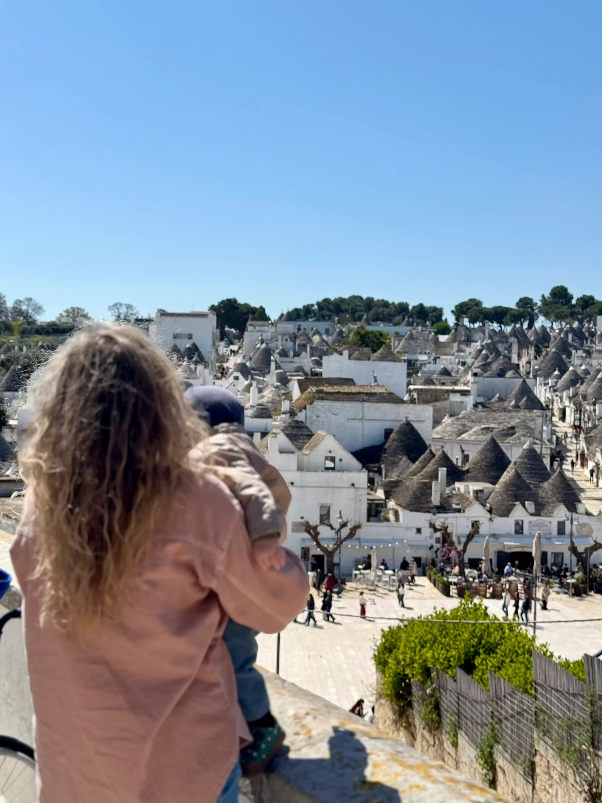 Garbanota moteris laiko kūdikį ant rankų ir žiūri į Alberobello trulli namų panoramą iš apžvalgos aikštelės
