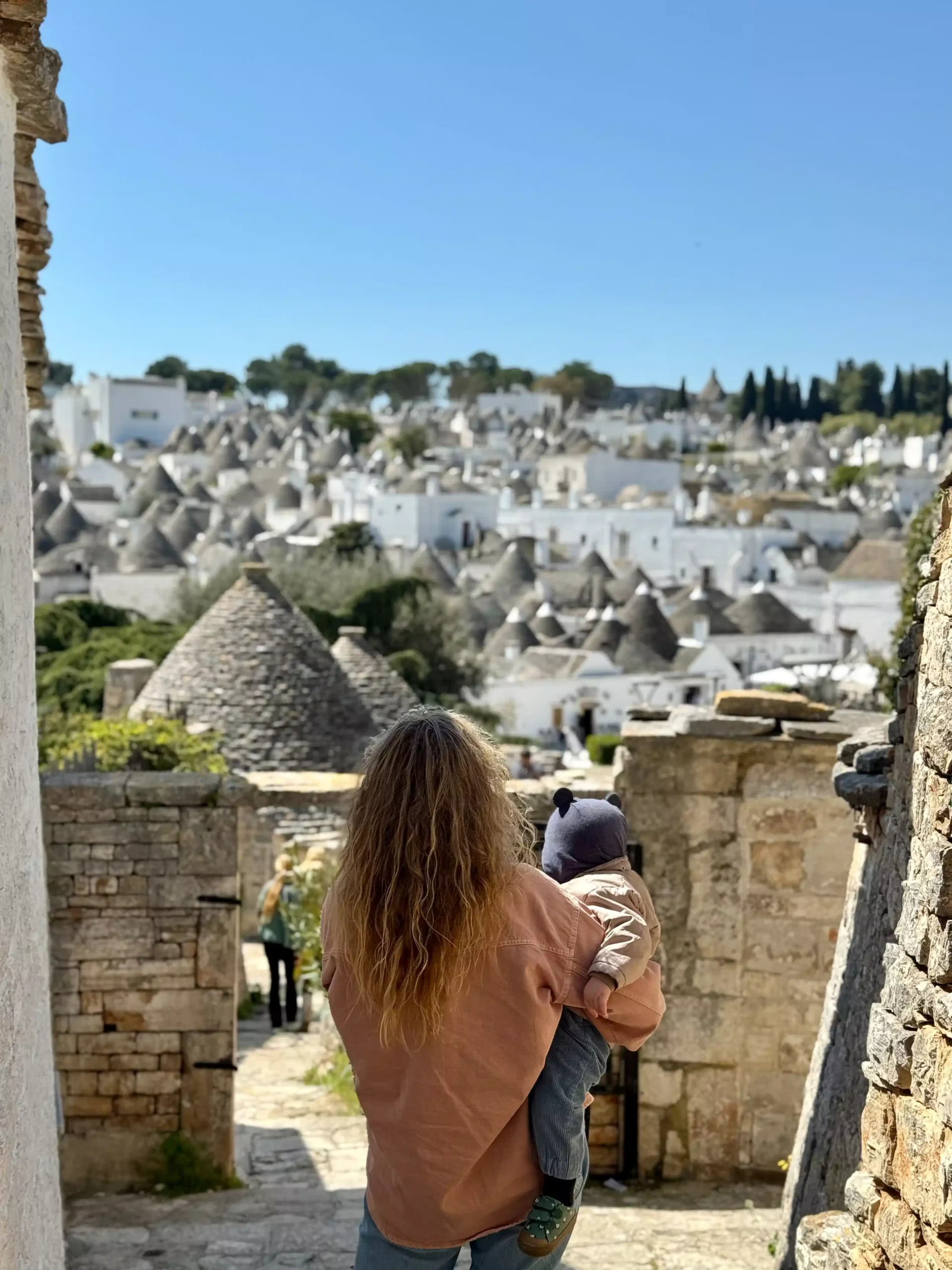 Moteris su mažu vaiku ant rankų stovi ant seno akmeninio tako ir stebi Alberobello trulli namelių miestelį iš aukštai