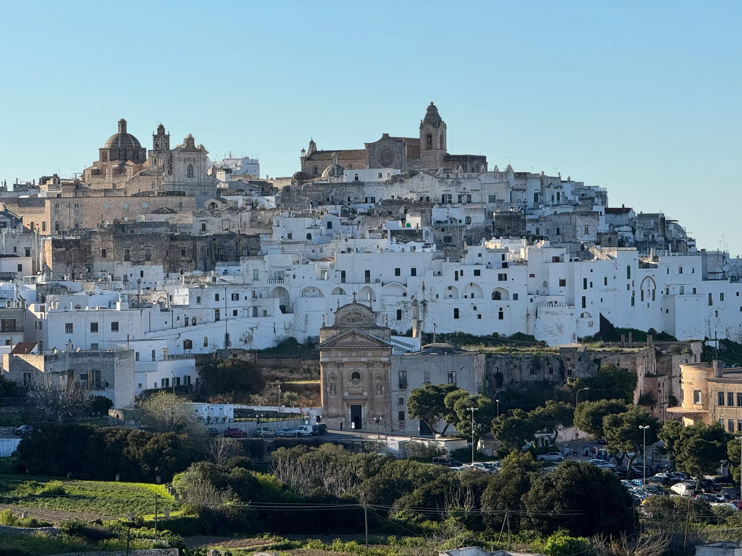Panoraminis vaizdas į Ostuni — vadinamąjį „baltąjį miestą" ant kalvos su bažnyčiomis ir tankiai sustatytais baltais namais, priešais žalios žemės sklypai