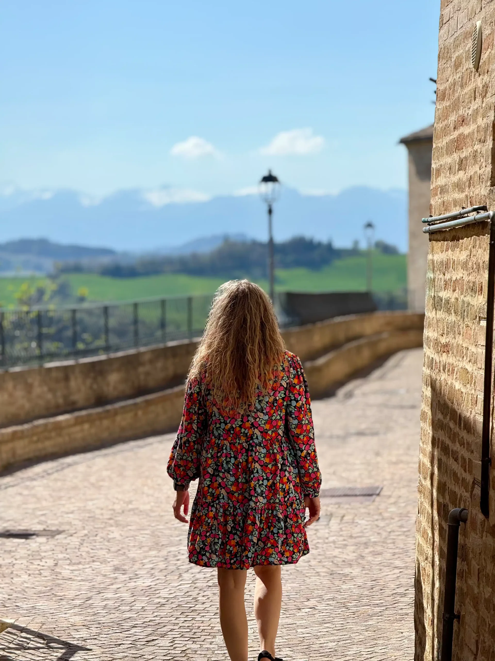 Marche su kūdikiu: Fermo ir slaptas Italijos perlas (2026) Moteris gėlėta suknele eina tolimo grindiniu grįstu alėju tarp akmeninių pastatų, fone žalios kalvos ir kalnai