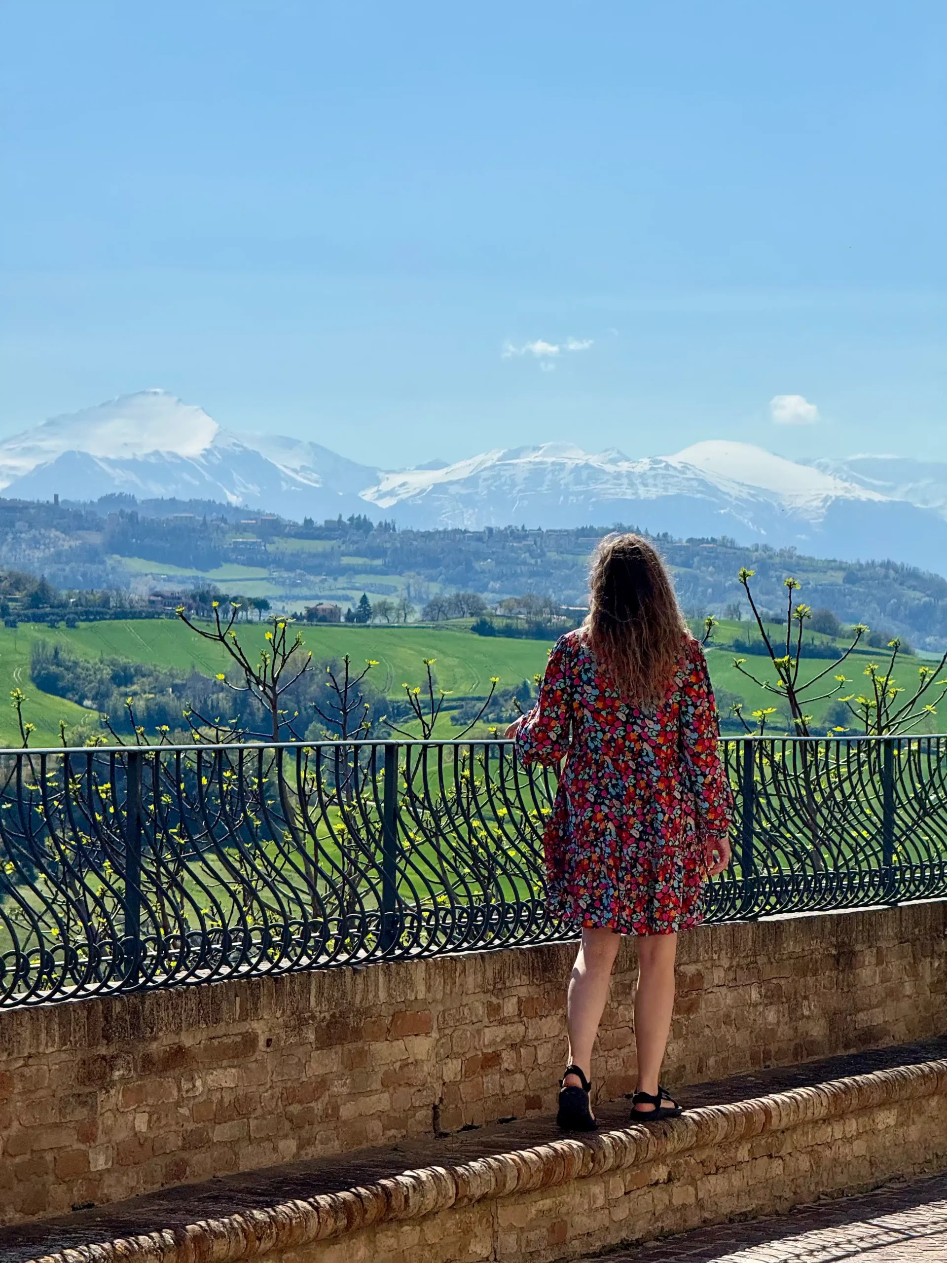 Marche su kūdikiu: Fermo ir slaptas Italijos perlas (2026) Moteris gėlėta suknele stovi ant akmeninio balkonėlio prie geležinės tvoros ir žvelgia į sniegais padengtas viršukalnes bei žaliuojančias Marche regiono kalvas