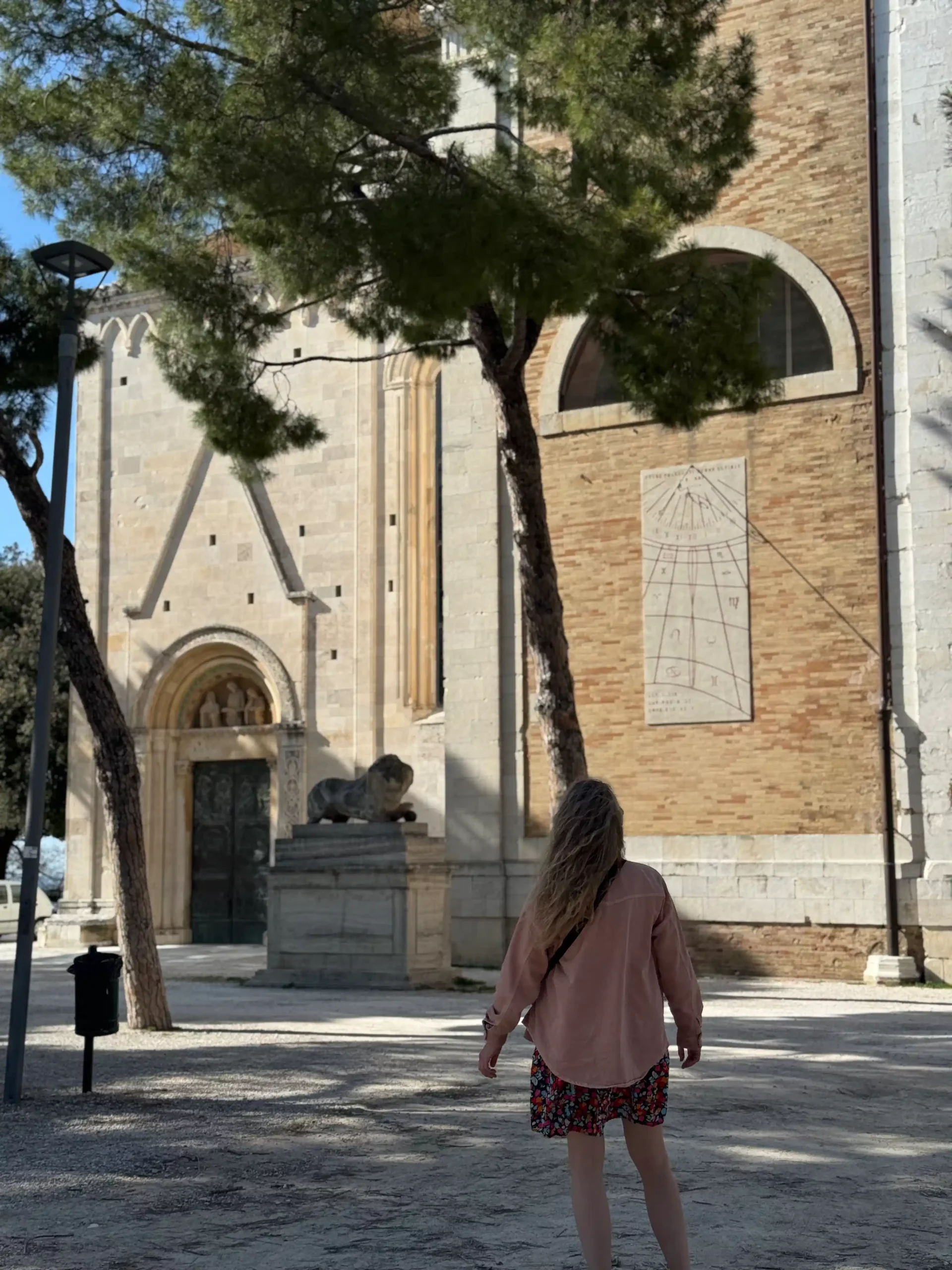 Marche su kūdikiu: Fermo ir slaptas Italijos perlas (2026) Moteris eina link viduramžių bažnyčios fasado su romaniniu portalu ir liūto skulptūra, ant sienos matomas saulės laikrodis, šalia – pušys